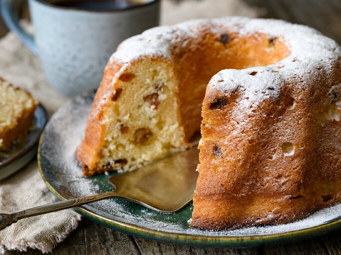 Frontalansicht: Ein angeschnittener Hefe-Gugelhupf steht auf einem Teller. Dahinter stehe eine Tasse und ein Stückchen Kuchen. | © Shutterstock / Stramp