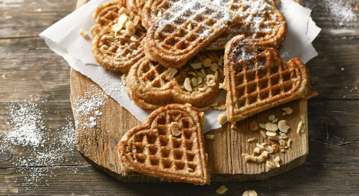 Waffeln in Herzform auf Krepp und hellbrauner Holzplatte angerichtet in der Draufsicht. Haselnusssplitter am Brettrand und den Waffeln verteil. Puderzucker darüber gestreut. Alles steht auf einem Holztisch mit dunkler Maserung. 