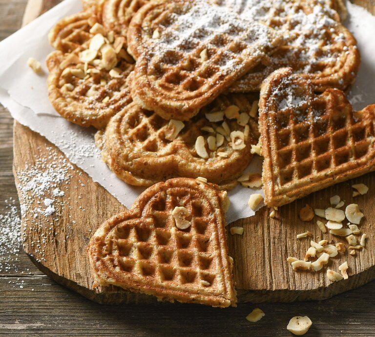 Waffeln in Herzform auf Krepp und hellbrauner Holzplatte angerichtet in der Draufsicht. Haselnusssplitter am Brettrand und den Waffeln verteil. Puderzucker darüber gestreut. Alles steht auf einem Holztisch mit dunkler Maserung.  | © StockFood / für ZS Verlag / Plewinski, Antje