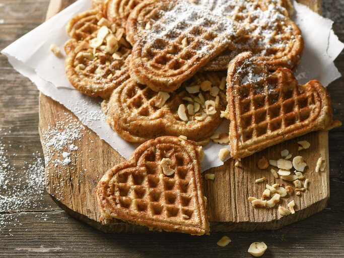 Waffeln in Herzform auf Krepp und hellbrauner Holzplatte angerichtet in der Draufsicht. Haselnusssplitter am Brettrand und den Waffeln verteil. Puderzucker darüber gestreut. Alles steht auf einem Holztisch mit dunkler Maserung.  | © StockFood / für ZS Verlag / Plewinski, Antje