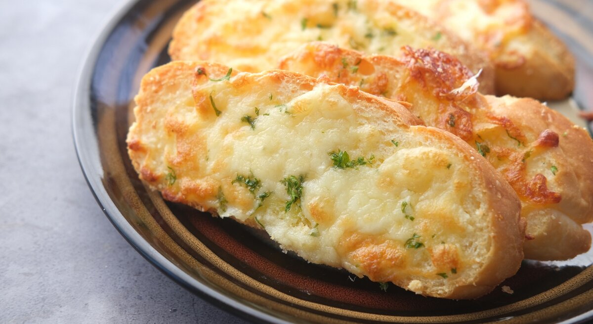 Käse-Knoblauch-Brot auf einem gemusterten Teller. Das Ganze auf einem grauen Untergrund. 