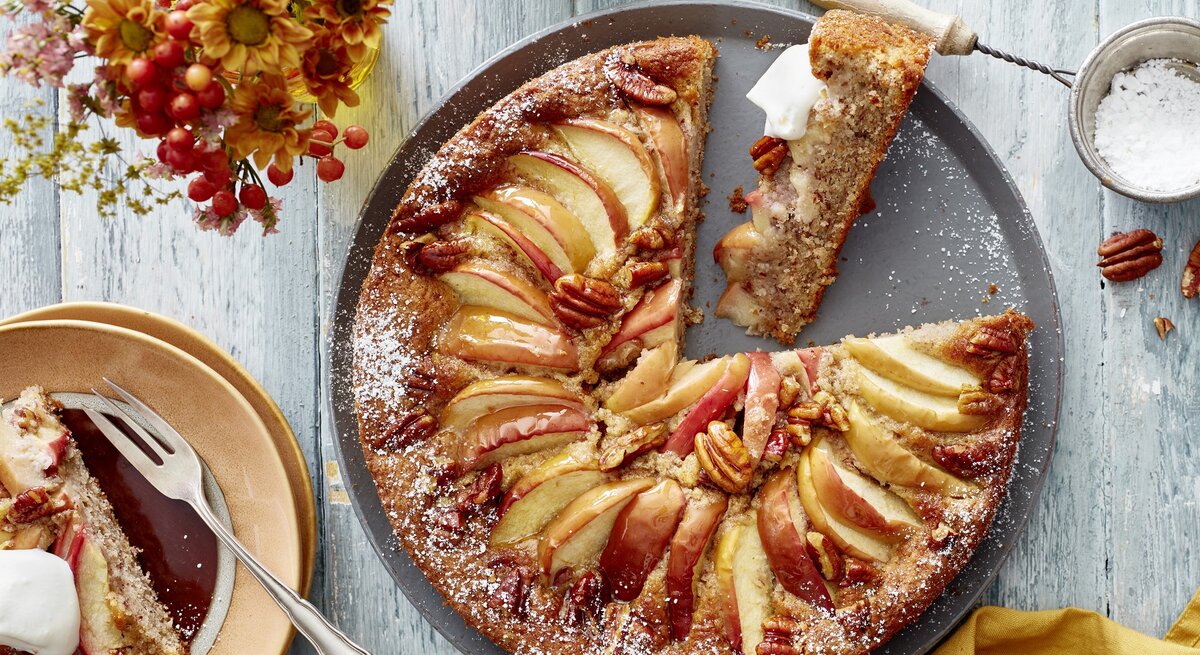 Ein grauer Teller mit dem Kuchen auf einem hellen Tisch direkt von oben fotografiert. Daneben rötliche Blumen und eine goldenes Tuch. 