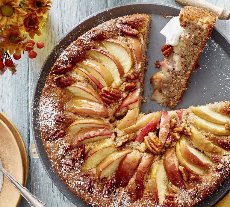 Ein grauer Teller mit dem Kuchen auf einem hellen Tisch direkt von oben fotografiert. Daneben rötliche Blumen und eine goldenes Tuch.  | © EAT CLUB