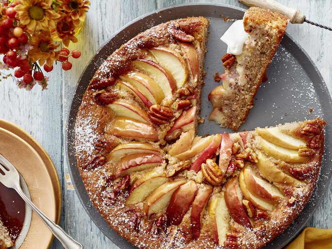 Ein grauer Teller mit dem Kuchen auf einem hellen Tisch direkt von oben fotografiert. Daneben rötliche Blumen und eine goldenes Tuch.  | © EAT CLUB