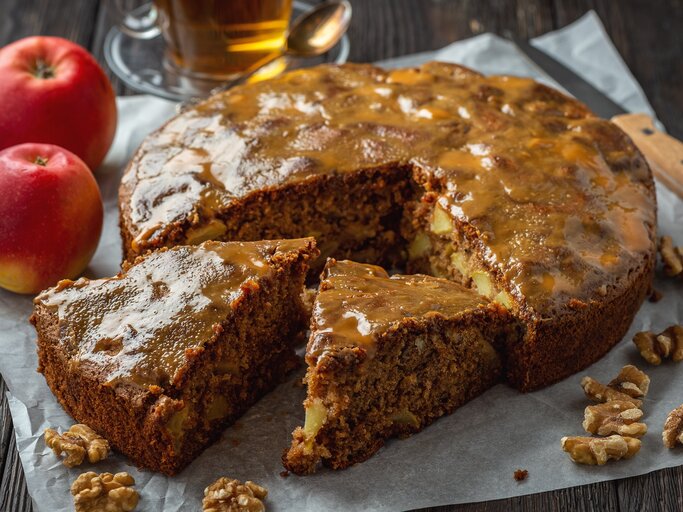 Glasierter Apfel-Walnusskuchen auf hölzernem Untergrund umgeben von den Hauptzutaten, einer Tasse und einem Messer.  | © Getty Images/iko636