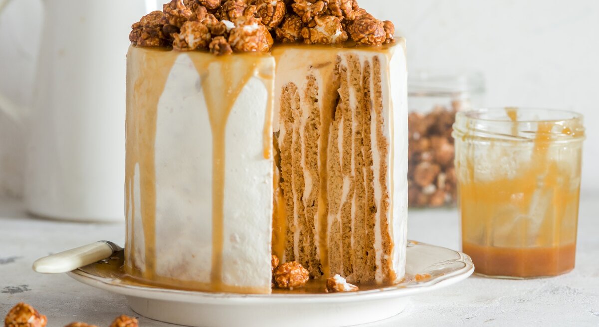 Angeschnittener Nusskuchen mit Karamell und Karamellpopcorn darauf, daneben Glas mit Karamell, heller Hintergrund. 