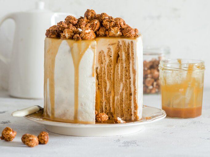 Angeschnittener Nusskuchen mit Karamell und Karamellpopcorn darauf, daneben Glas mit Karamell, heller Hintergrund.  | © Stockfood / Meliukh Irina
