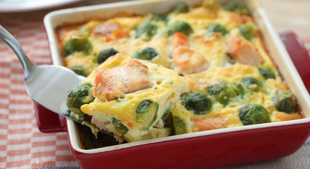 Ein Rosenkohl-Lachs-Gratin in einer weiß-roten Form steht auf einem gemusterten Geschirrtuch. Das Gratin ist bereits angeschnitten und ein Stück wird mit einem Tortenheber herausgehoben.