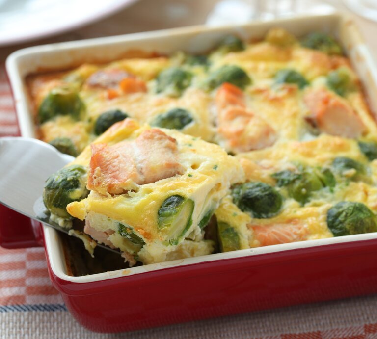 Ein Rosenkohl-Lachs-Gratin in einer weiß-roten Form steht auf einem gemusterten Geschirrtuch. Das Gratin ist bereits angeschnitten und ein Stück wird mit einem Tortenheber herausgehoben. | © Shutterstock / StockphotoVideo