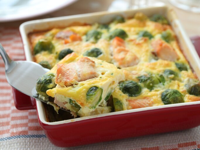 Ein Rosenkohl-Lachs-Gratin in einer weiß-roten Form steht auf einem gemusterten Geschirrtuch. Das Gratin ist bereits angeschnitten und ein Stück wird mit einem Tortenheber herausgehoben. | © Shutterstock / StockphotoVideo