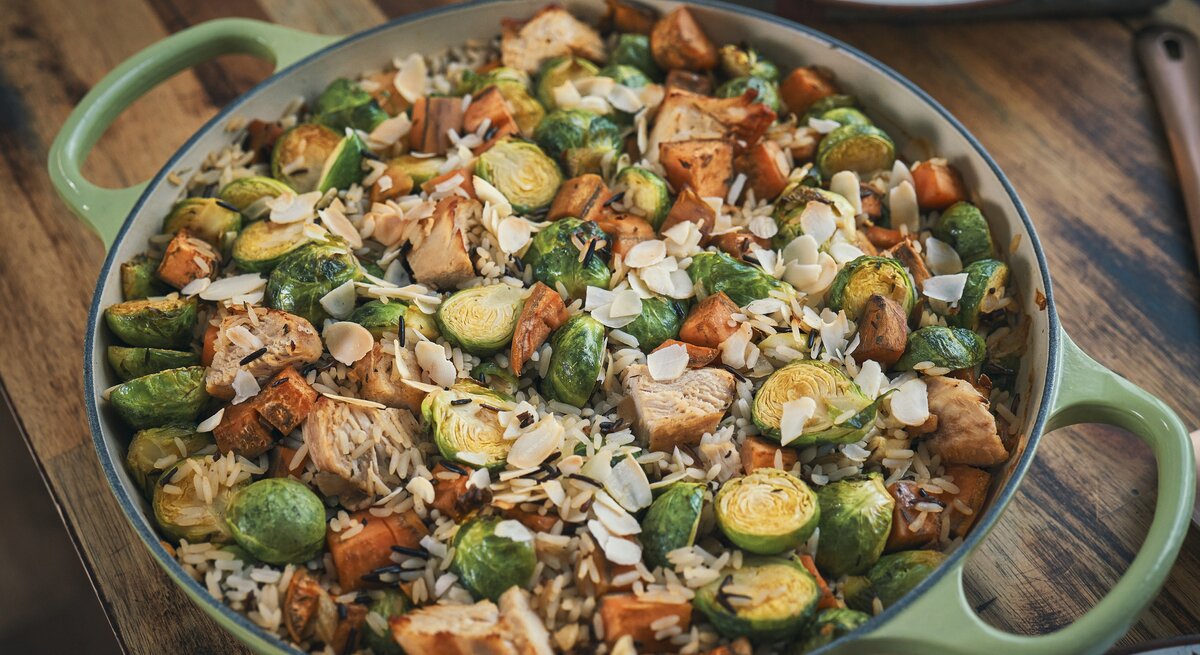 Eine Rosenkohl-Huhn-Reis-Pfanne in einer großen, grünen Pfanne mit hohem Rand. Die Reispfanne ist angerichtet mit Mandelblättchen und steht auf einem braunen Holztisch.