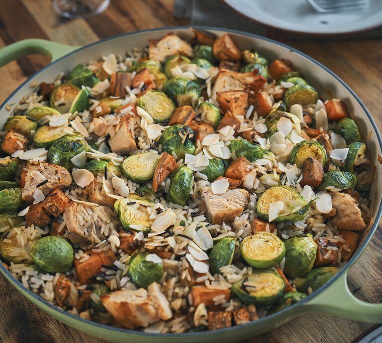 Eine Rosenkohl-Huhn-Reis-Pfanne in einer großen, grünen Pfanne mit hohem Rand. Die Reispfanne ist angerichtet mit Mandelblättchen und steht auf einem braunen Holztisch. | © Getty Images / GMVozd