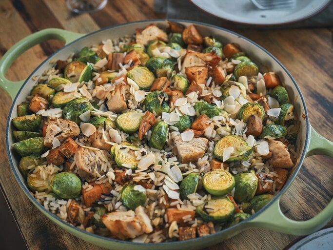 Eine Rosenkohl-Huhn-Reis-Pfanne in einer großen, grünen Pfanne mit hohem Rand. Die Reispfanne ist angerichtet mit Mandelblättchen und steht auf einem braunen Holztisch. | © Getty Images / GMVozd