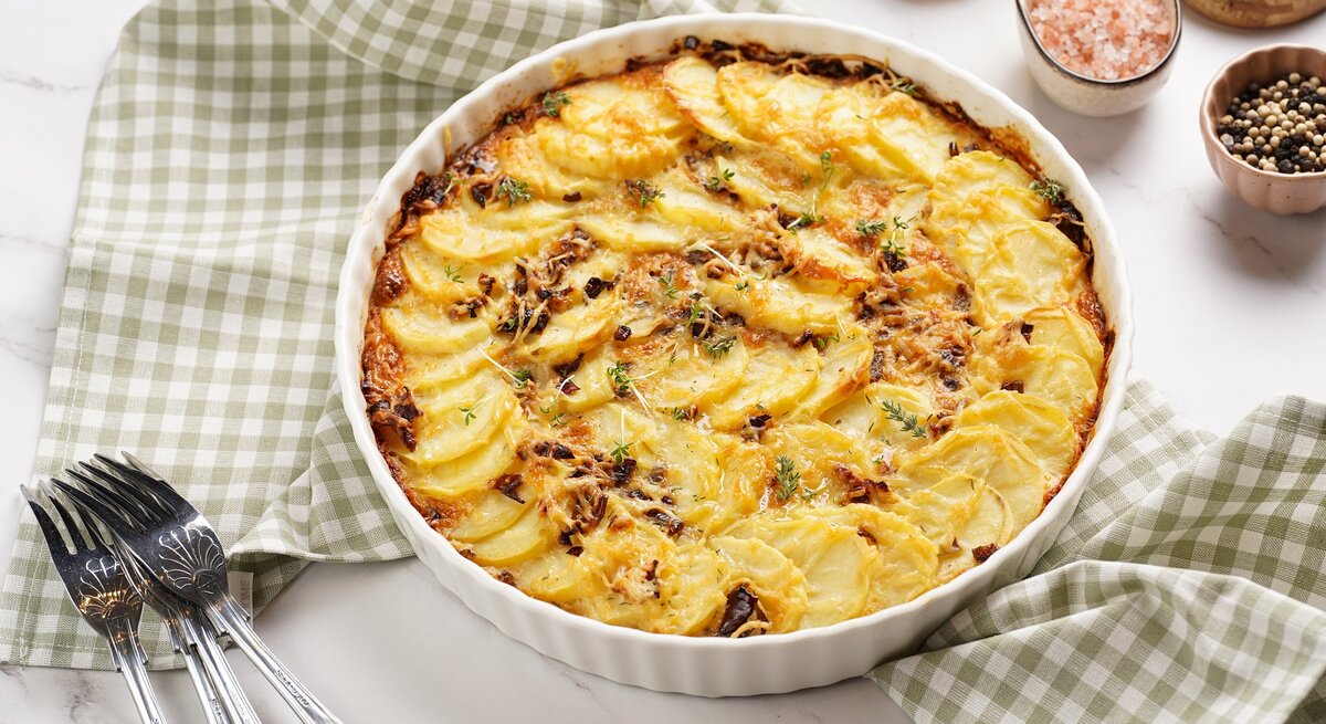 Eine runde weiße Auflaufform mit dem Kartoffelgratin auf einem weißen Tisch mit grün-weiß-karierter Tischdecke.