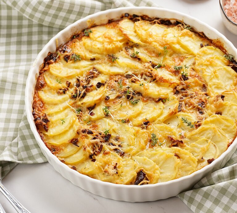 Eine runde weiße Auflaufform mit dem Kartoffelgratin auf einem weißen Tisch mit grün-weiß-karierter Tischdecke. | © Shutterstock/ RomanaMart 