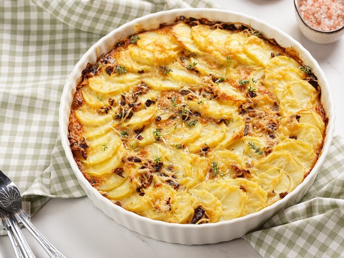 Eine runde weiße Auflaufform mit dem Kartoffelgratin auf einem weißen Tisch mit grün-weiß-karierter Tischdecke. | © Shutterstock/ RomanaMart 