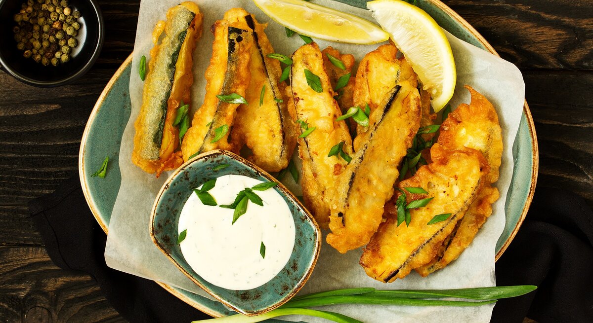 Draufsicht: asiatische Zucchini-Sticks die in einer Pfanne frittiert wurden. Daneben liegen zwei Zitronenschnitzer, eine Schale mit Pfefferkörnern und ein Schale mit Joghurt-Dip.