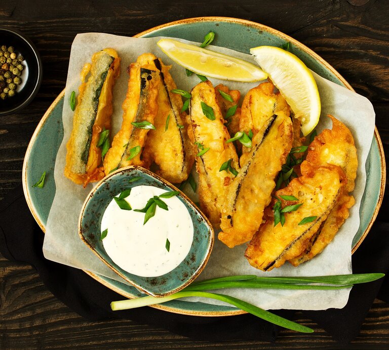 Draufsicht: asiatische Zucchini-Sticks die in einer Pfanne frittiert wurden. Daneben liegen zwei Zitronenschnitzer, eine Schale mit Pfefferkörnern und ein Schale mit Joghurt-Dip. | © Shutterstock / Lesya Dolyuk