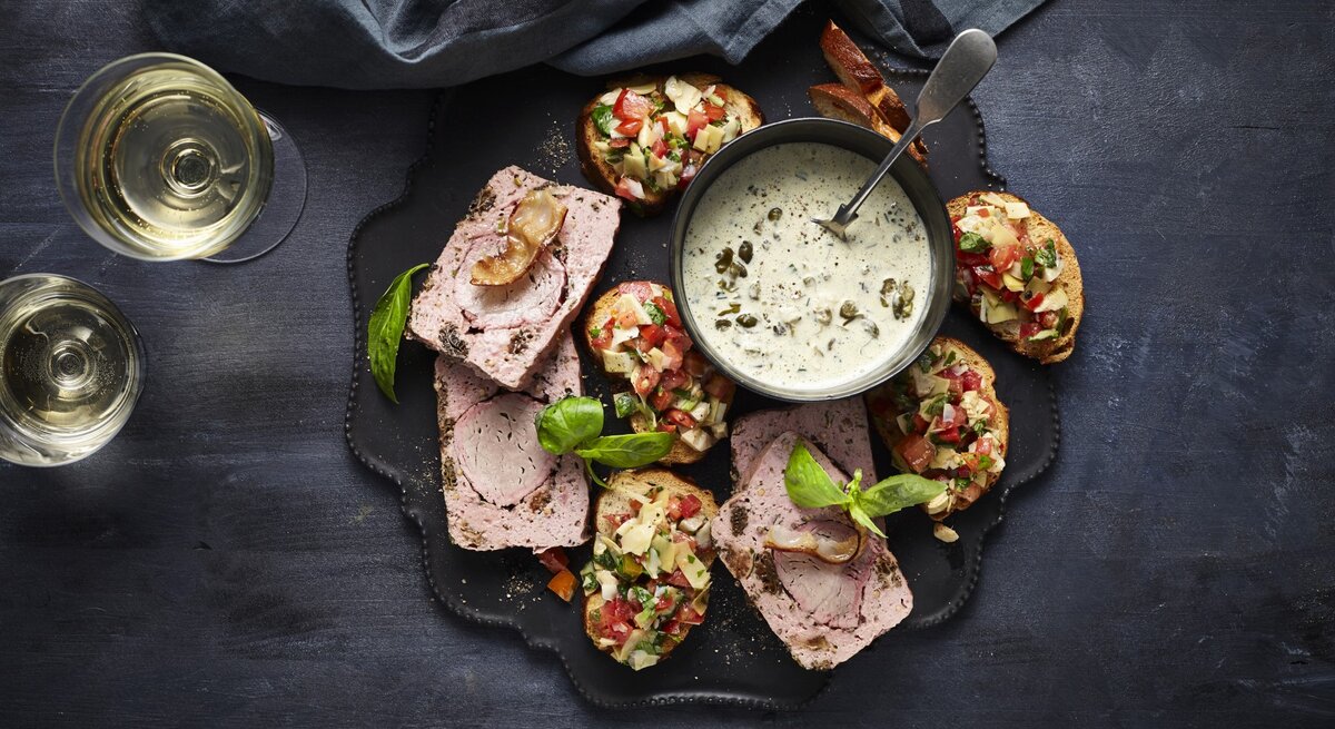 Leberkäse de luxe mit Laugen-Crostini und Artischocken-Salsa auf einer Snackplatte. Daneben zwei Gläser Sekt und ein dunkles Küchentuch. 