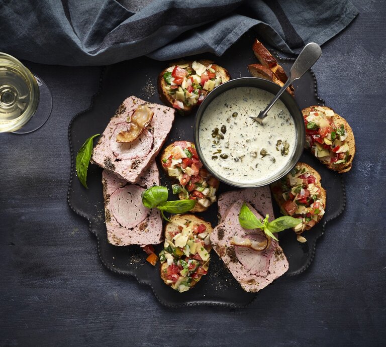 Leberkäse de luxe mit Laugen-Crostini und Artischocken-Salsa auf einer Snackplatte. Daneben zwei Gläser Sekt und ein dunkles Küchentuch.  | © StockFood / StockFood Studios / Holsten, Ulrike