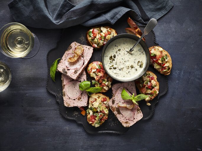 Leberkäse de luxe mit Laugen-Crostini und Artischocken-Salsa auf einer Snackplatte. Daneben zwei Gläser Sekt und ein dunkles Küchentuch.  | © StockFood / StockFood Studios / Holsten, Ulrike