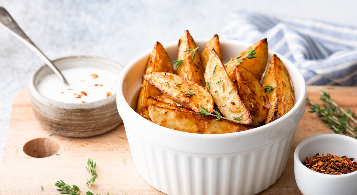Kohlrabi-Wedges mit Thymian in einer weißen Schale auf einem Brett, darauf außerdem eine Schale Dip und eine Schale mit getrockneten Gewürzen. 
