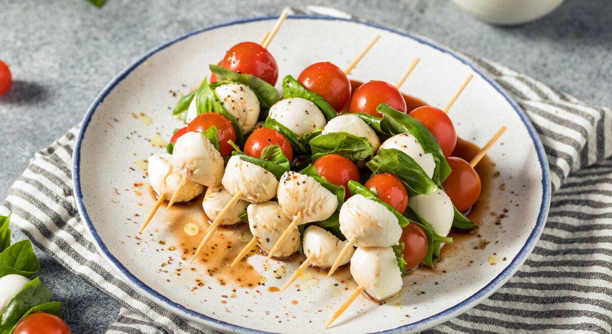 Fünf Tomaten-Mozzarella-Spieße liegen auf einem Teller. Neben dem Teller liegen einzelne Kirschtomaten und ein paar Blätter Basilikum.