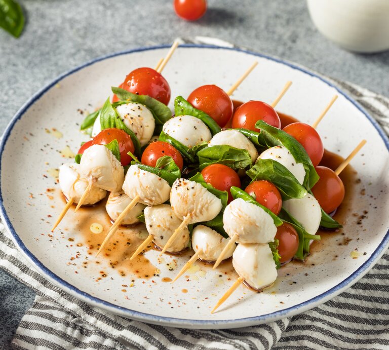 Fünf Tomaten-Mozzarella-Spieße liegen auf einem Teller. Neben dem Teller liegen einzelne Kirschtomaten und ein paar Blätter Basilikum. | © Getty Images / 	bhofack2