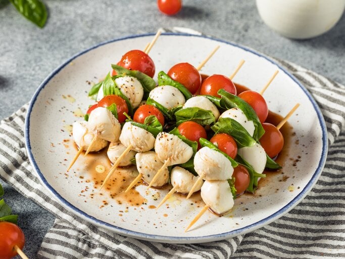 Fünf Tomaten-Mozzarella-Spieße liegen auf einem Teller. Neben dem Teller liegen einzelne Kirschtomaten und ein paar Blätter Basilikum. | © Getty Images / 	bhofack2