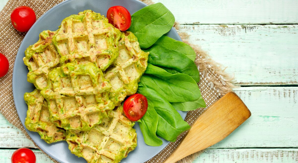 Draufsicht: helle Holzplanken auf denen ein grobes Leintuch liegt. Auf dem ist ein blauer Teller mit Falafel-Waffeln mit Spinat, Daneben liegen frische Spinatblätter und Kirschtomaten.