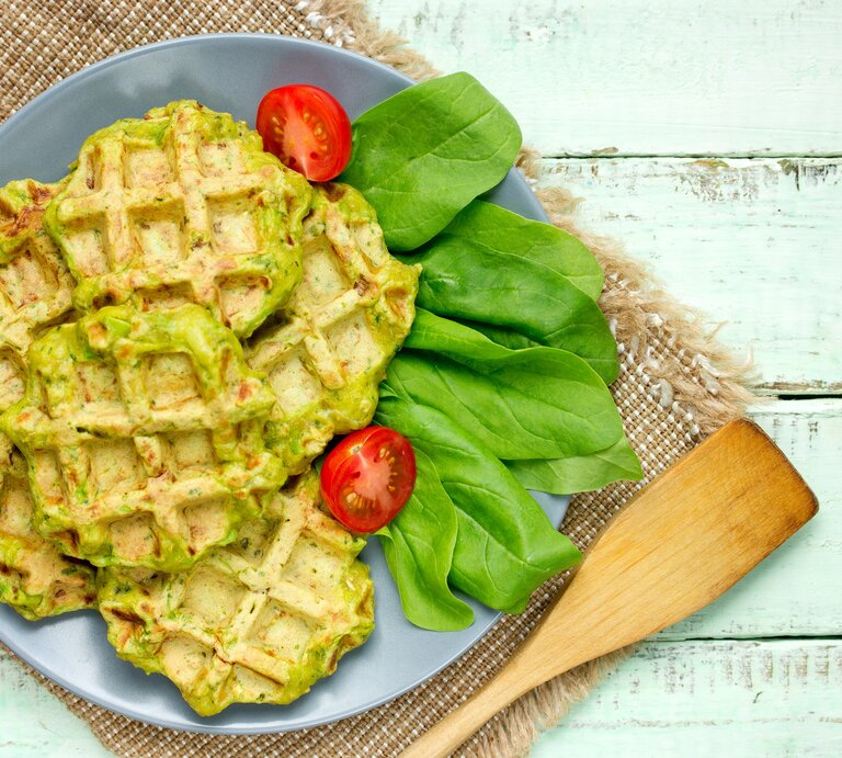Draufsicht: helle Holzplanken auf denen ein grobes Leintuch liegt. Auf dem ist ein blauer Teller mit Falafel-Waffeln mit Spinat, Daneben liegen frische Spinatblätter und Kirschtomaten. | © Shutterstock / Anastasia_Panait