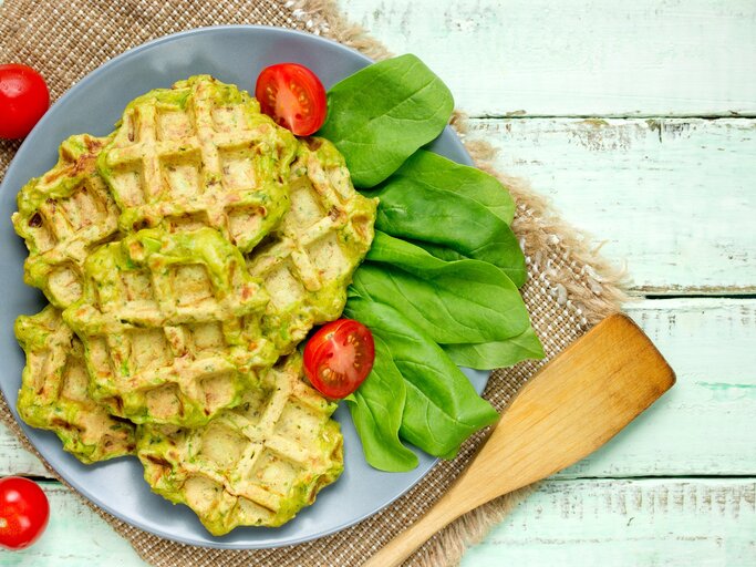 Draufsicht: helle Holzplanken auf denen ein grobes Leintuch liegt. Auf dem ist ein blauer Teller mit Falafel-Waffeln mit Spinat, Daneben liegen frische Spinatblätter und Kirschtomaten. | © Shutterstock / Anastasia_Panait