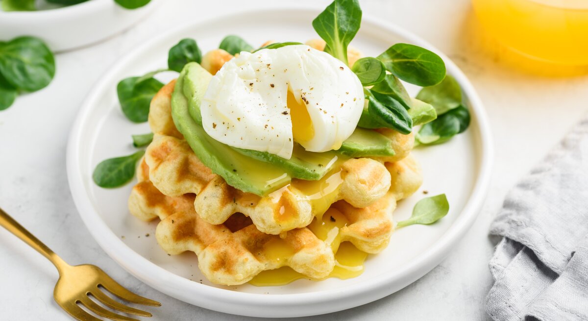 Käsewaffel mit Avocadoscheiben und pochiertem Ei oben, auf einem weißen Teller mit ein paar Feldsalatblättern garniert