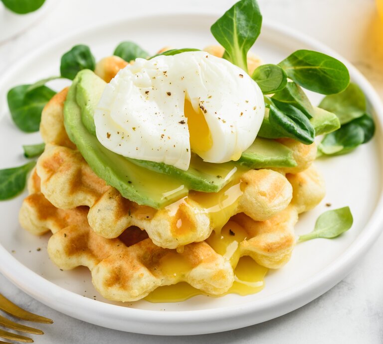 Käsewaffel mit Avocadoscheiben und pochiertem Ei oben, auf einem weißen Teller mit ein paar Feldsalatblättern garniert | © Shutterstock/rom_olik
