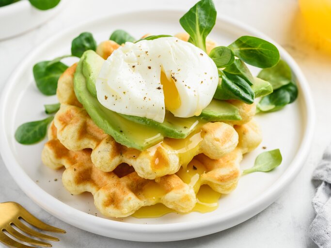 Käsewaffel mit Avocadoscheiben und pochiertem Ei oben, auf einem weißen Teller mit ein paar Feldsalatblättern garniert | © Shutterstock/rom_olik