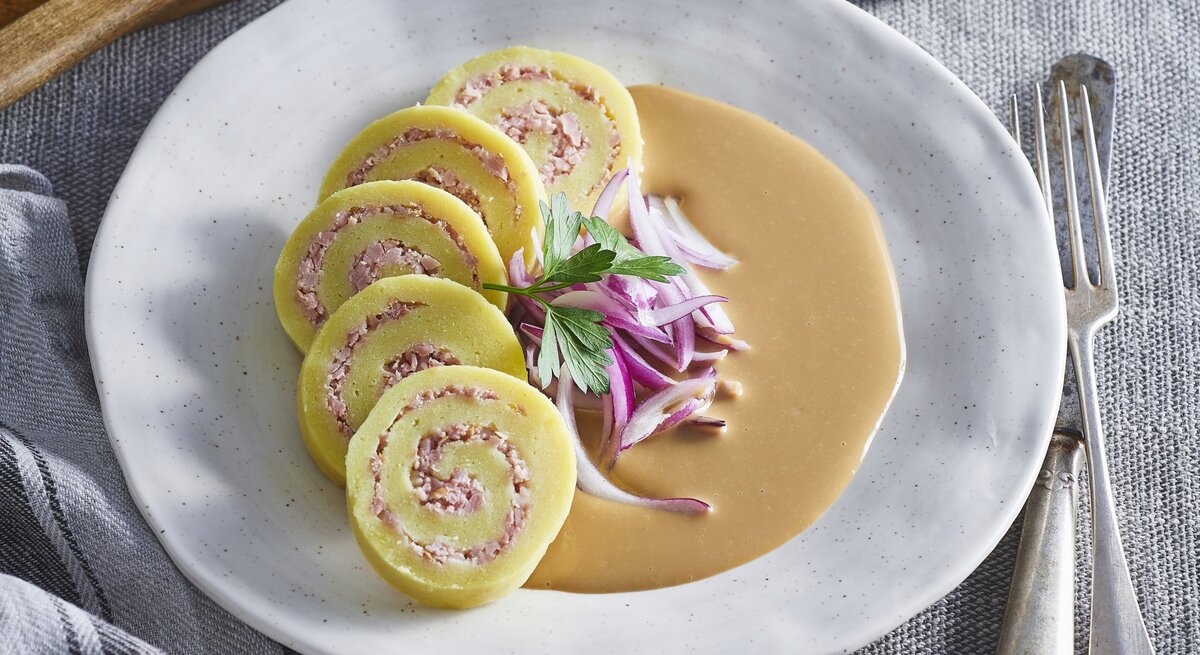 Kartoffelrouladen mit Schweinefleischfüllung auf Zwiebelsauce auf einem weißen Teller. Daneben liegt Besteck. 