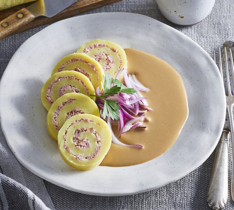 Kartoffelrouladen mit Schweinefleischfüllung auf Zwiebelsauce auf einem weißen Teller. Daneben liegt Besteck.  | ©  StockFood / Profimedia