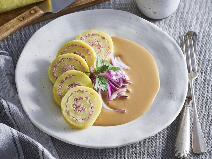 Kartoffelrouladen mit Schweinefleischfüllung auf Zwiebelsauce auf einem weißen Teller. Daneben liegt Besteck.  | ©  StockFood / Profimedia