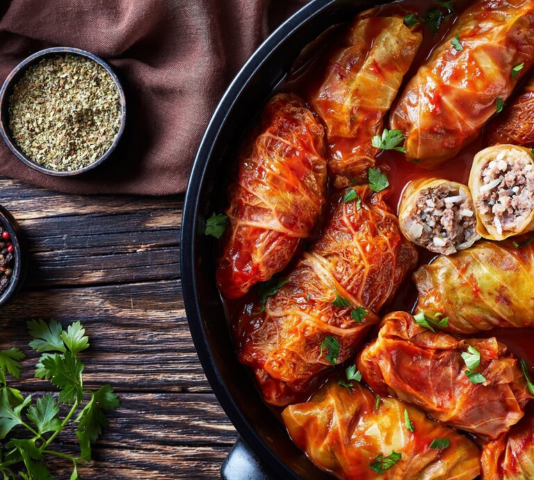 Eine schwarze Pfanne mit Kohlrouladen in Tomatensoße. Daneben Gewürze und Petersilie. Alles von oben auf einem dunklen Holztisch fotografiert. | © Shutterstock/ from my point of view