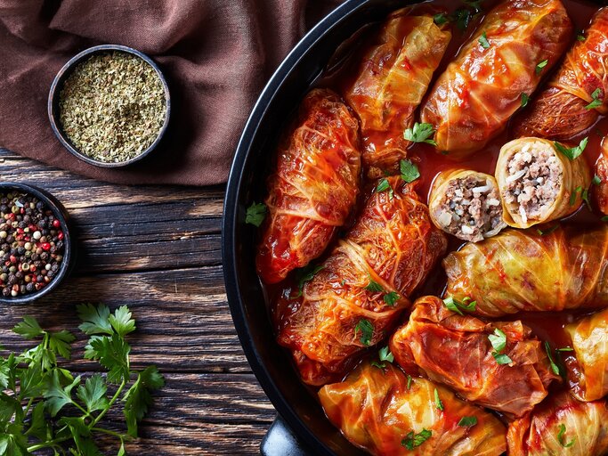 Eine schwarze Pfanne mit Kohlrouladen in Tomatensoße. Daneben Gewürze und Petersilie. Alles von oben auf einem dunklen Holztisch fotografiert. | © Shutterstock/ from my point of view