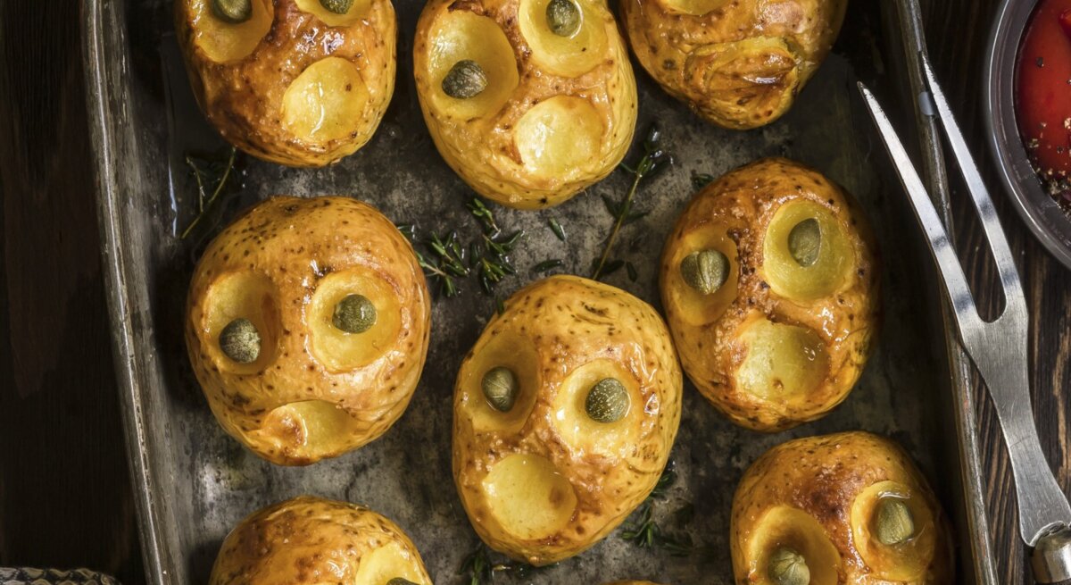 Halloween-Bratkartoffeln mit Kapernaugen, geschnitzt mit gruseligen Fratzen und serviert auf einem schwarzen Blech auf hölzernem Untergrund. 