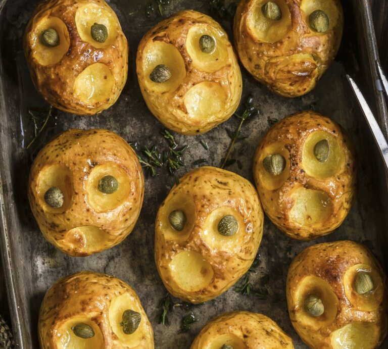 Halloween-Bratkartoffeln mit Kapernaugen, geschnitzt mit gruseligen Fratzen und serviert auf einem schwarzen Blech auf hölzernem Untergrund.  | © StockFood / Tatiana, Vorona