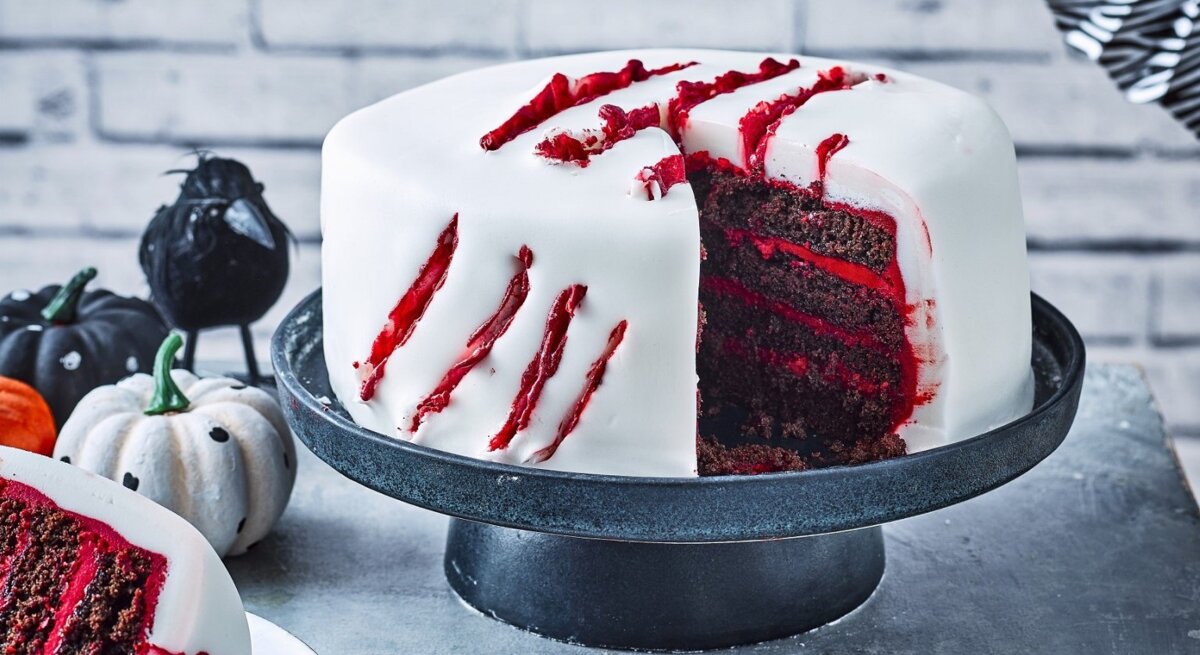 Auf einem Tortenteller steht eine gruselige Halloween-Torte. Sie ist mit weißem Fondant eingedeckt und hat blutige Risse als Dekoration.
