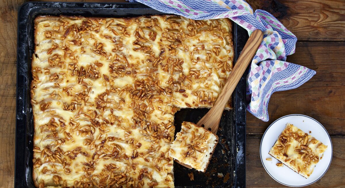 Apfelkuchen vom Blech mit Krokant, angeschnitten auf dem schwarzen Ofenblech serviert. 