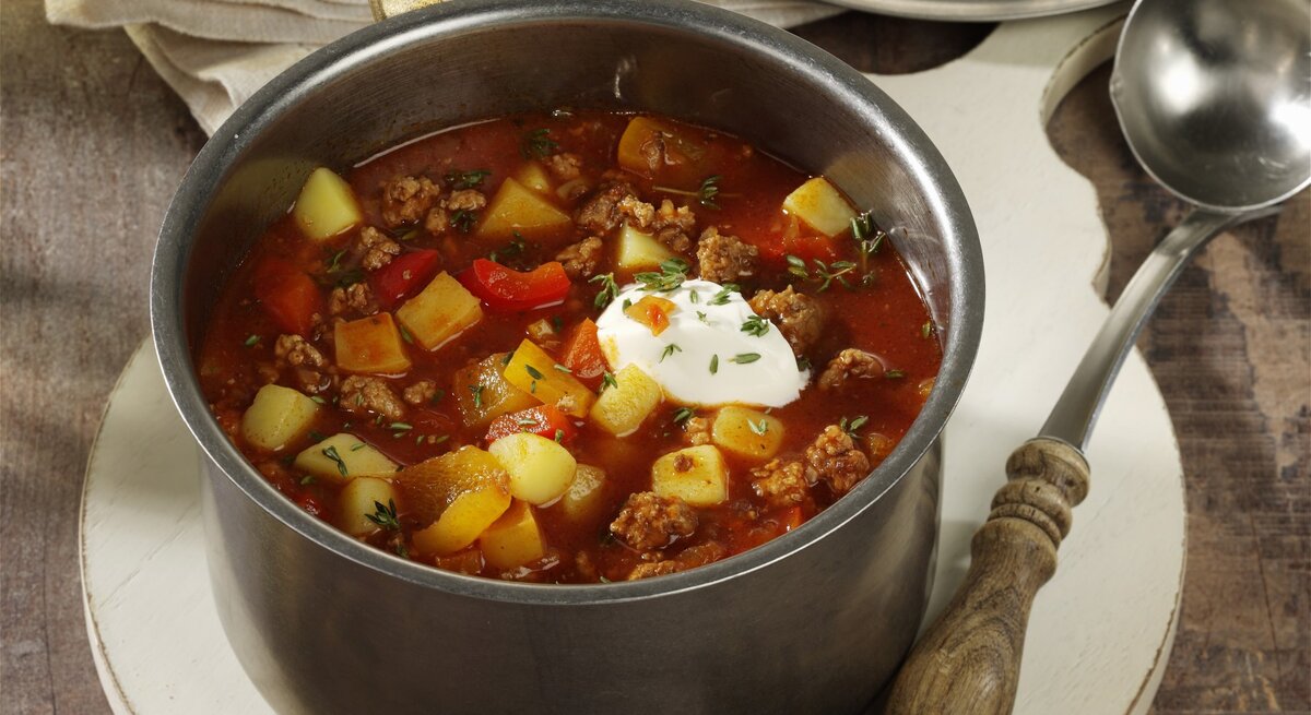 Bauerneintopf mit Hackfleisch, Paprika ud Kartoffeln, garniert mit Schmand und frischem Schnittlauch, serviert in einem großen Topf auf einem rustikalen Holztisch. 