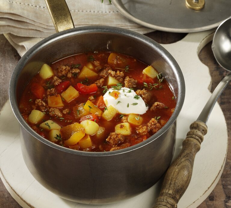 Bauerneintopf mit Hackfleisch, Paprika ud Kartoffeln, garniert mit Schmand und frischem Schnittlauch, serviert in einem großen Topf auf einem rustikalen Holztisch.  | © StockFood / StockFood Studios / Photoart