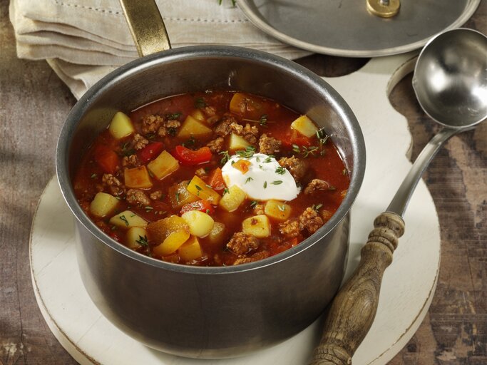 Bauerneintopf mit Hackfleisch, Paprika ud Kartoffeln, garniert mit Schmand und frischem Schnittlauch, serviert in einem großen Topf auf einem rustikalen Holztisch.  | © StockFood / StockFood Studios / Photoart