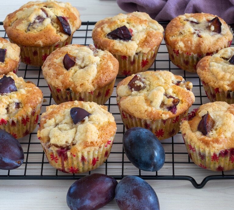 Pflaumen-Muffins mit griechischem Joghurt liegen auf einem Gitter. Daneben liegt ein violettes Tuch und einige Pflaumen.  | © Getty Images / 	AndreaRR