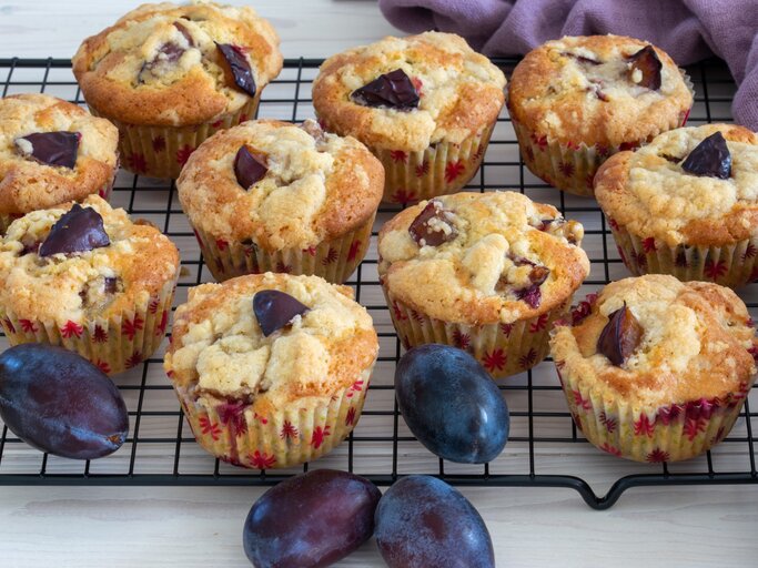 Pflaumen-Muffins mit griechischem Joghurt liegen auf einem Gitter. Daneben liegt ein violettes Tuch und einige Pflaumen.  | © Getty Images / 	AndreaRR