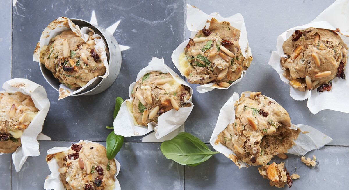 Vegetarische Gemüse-Pizza-Muffins mit Pinienkernen und frischem Basilikum, eingewickelt in Backpapier und drappiert auf einem hellen Metalltisch. 